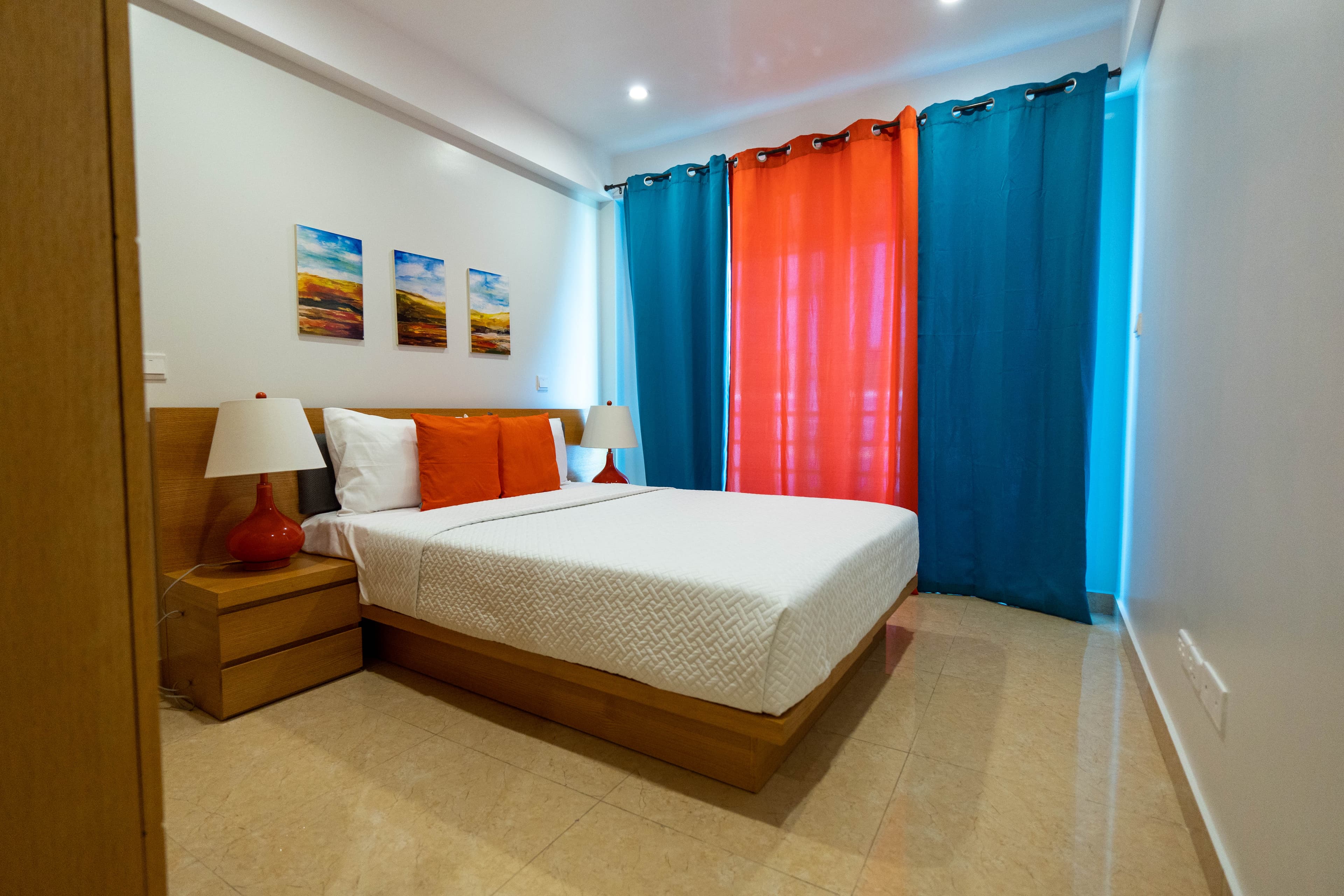 Master bedroom with queen platform bed, wooden nightstand with lamp, and floor to ceiling windows with sunset orange and ocean blue drapes