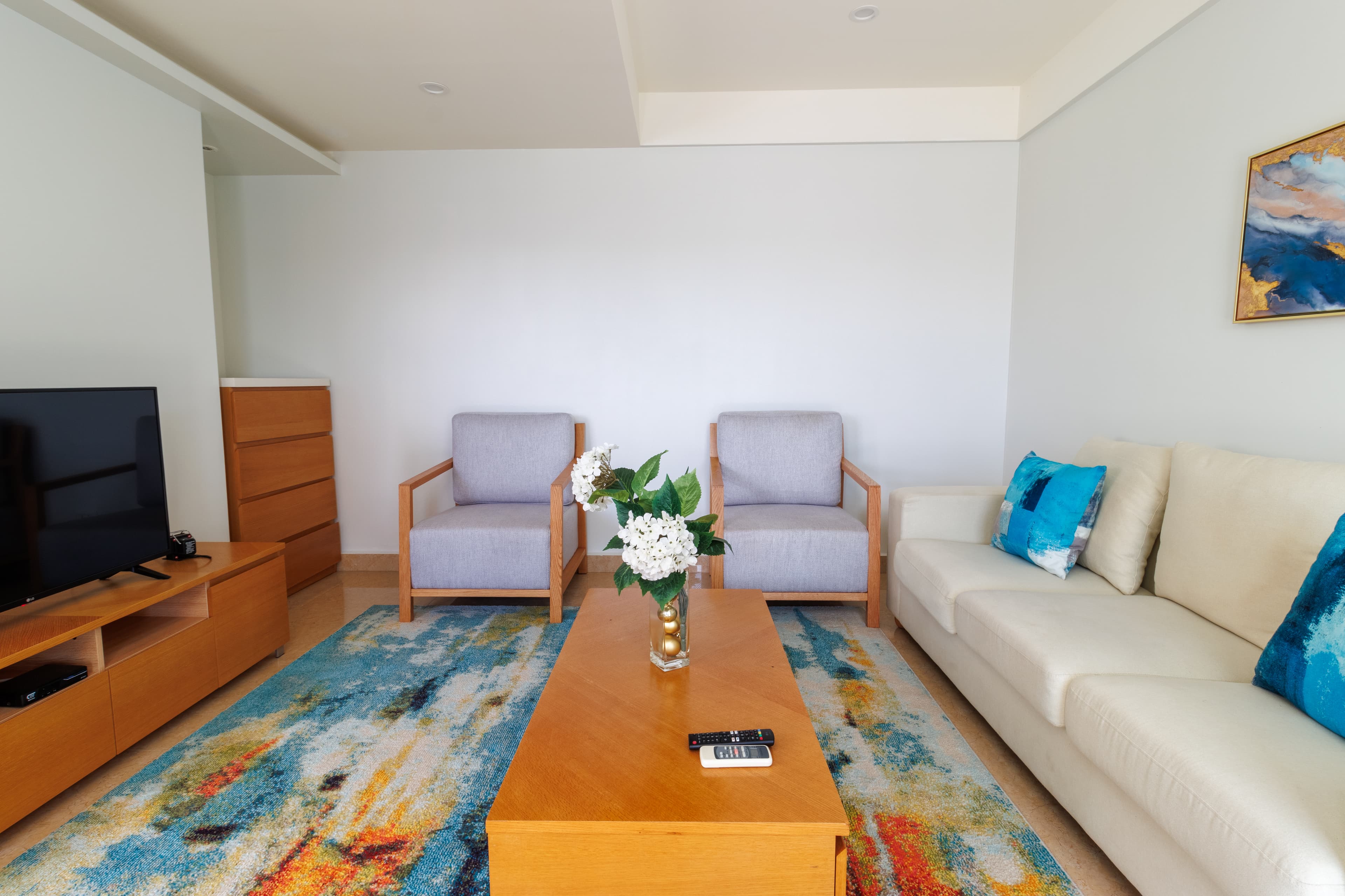 Tranquil living room with long cream colored upholstered couch, two upholstered armchairs, wooden coffee table on a modern area rug & console table with flat-screen TV