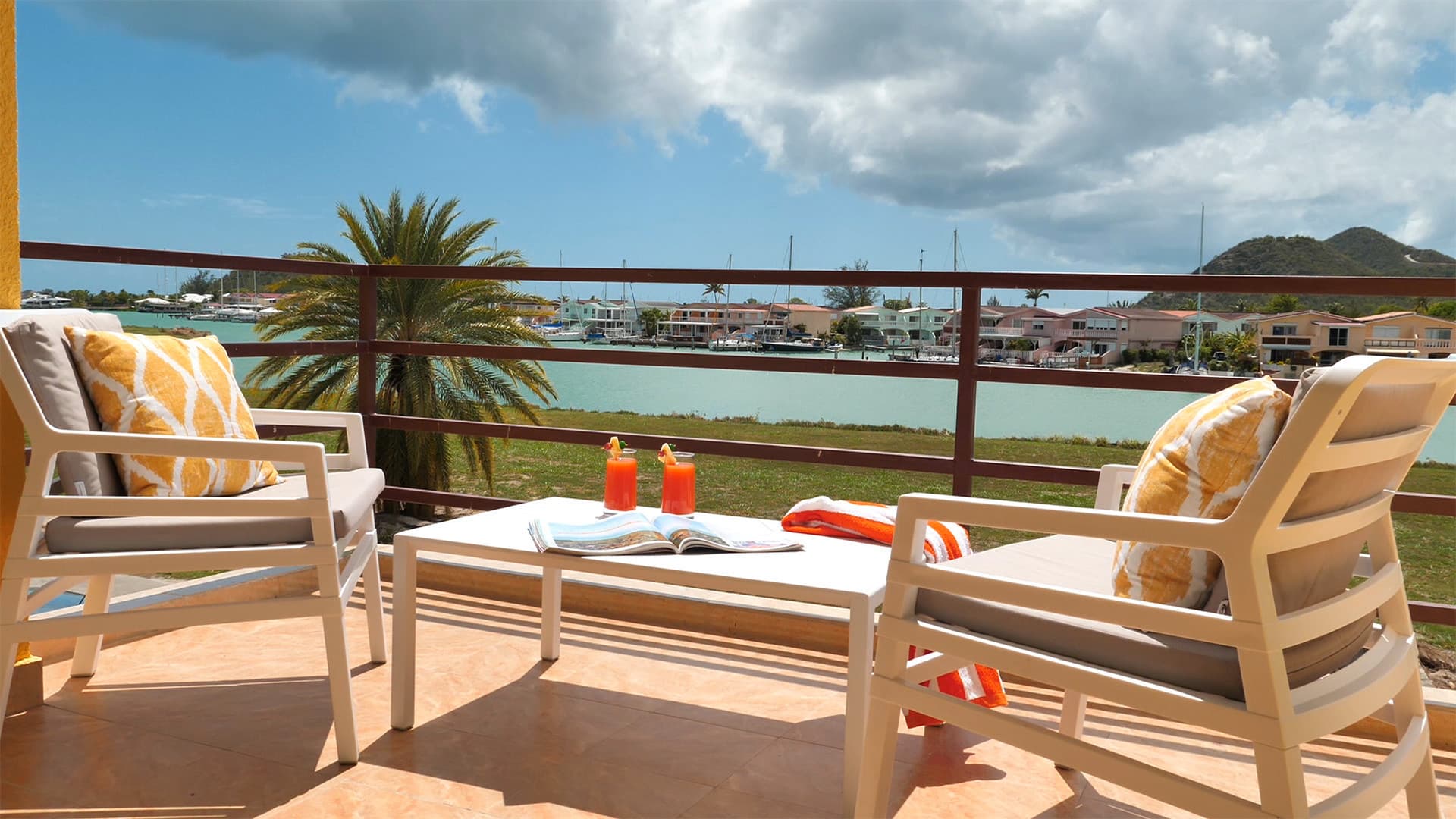 Two lounge chairs with fresh juice in glasses on table over looking nice view of boats in a harbor
