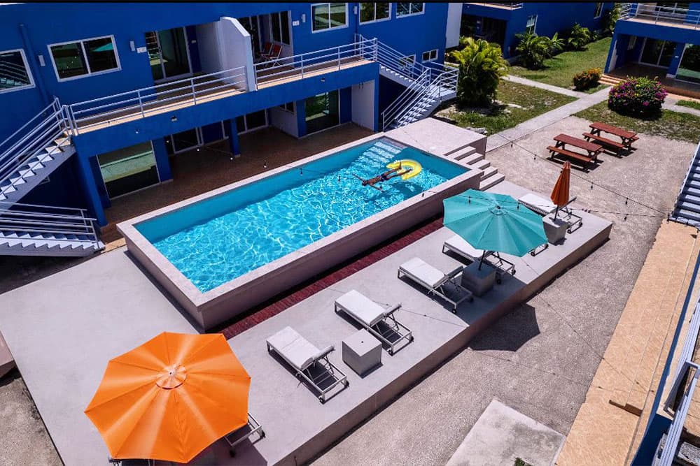 A rectangular swimming pool surrounded by lounge chairs and umbrellas. The pool has clear blue water with a person floating in an inflatable tube. There are buildings with balconies in the background, and lush greenery.